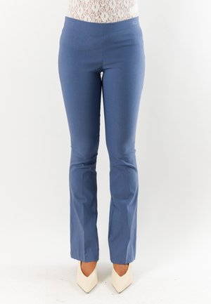 Donna con pantaloni blu aderenti a gamba dritta e tacchi alti bianchi appuntiti, in piedi davanti a uno sfondo chiaro e uniforme.