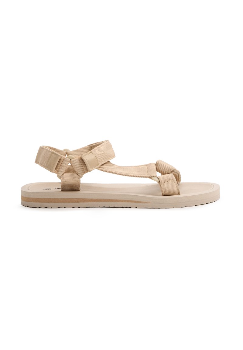 Sandalias planas beige con suela de goma texturizada, con tiras de tela ajustables y un diseño sencillo. Ligeras y prácticas para uso al aire libre.