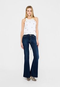 Bela brezrokavnik z okrasnimi zlati poudarki, kombiniran z razširjenimi temno modrimi kavbojkami in vzorčastimi gležnarji. Enakomerna tekstura po celoti.