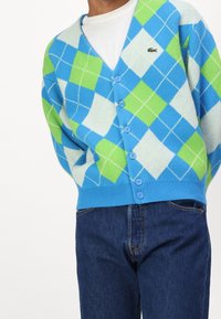 Hombre con un cardigans de rombos azul, verde y blanco abrochado sobre una camisa blanca, combinado con jeans de mezclilla azul oscuro, mostrado de los hombros a la cintura.