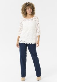 Top bianco in pizzo con maniche a tre quarti, motivo floreale, orlo a scallop; abbinato a jeans blu scuro e stivaletti beige.