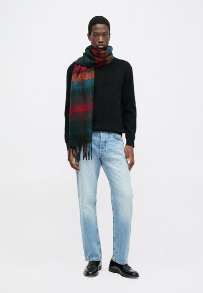 Giovane uomo che indossa un maglione nero, una sciarpa a scacchi multicolore, jeans azzurri e scarpe eleganti nere, in piedi contro uno sfondo bianco semplice.
