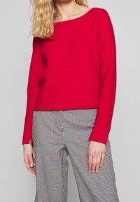 Suéter texturizado rojo con escote amplio, mangas largas y puños acanalados, combinado con pantalones de pierna ancha a cuadros en blanco y negro.
