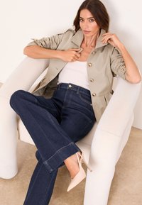 Naine istub valges tugitoolis, kannab beežikat nööbitud jopet, valget topi, tumedaid laia säärega teksapükse ja värvitu tugeva kontsaga kingi.