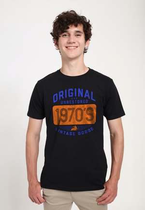 Jonge man met krullend donker haar die glimlacht, gekleed in een zwart T-shirt met blauwe en oranje tekst waarop staat "Original and Unrestored 1970's Vintage Goods."