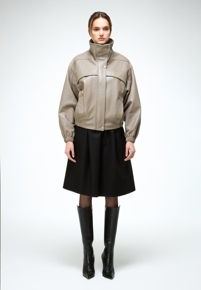 Frau trägt taupefarbenen Lederjacke mit hohem Kragen, schwarzen A-Linien-Rock, schwarze Strumpfhose und schwarze kniehohe Stiefel, steht frontal vor weißem Hintergrund.