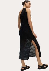Robe en crochet noire sans manches avec des motifs verticaux et horizontaux, dotée d'une fente sur le côté et d'une longueur mi-mollet. Assortie à des sandales noires.