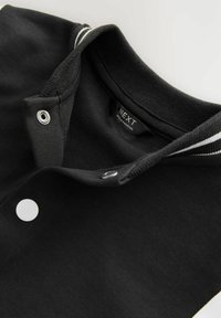Cuello de chaqueta varsity negra con ribete de rayas blancas y dos botones de presión visibles, mostrando la etiqueta de la marca "NEXT" en el interior.