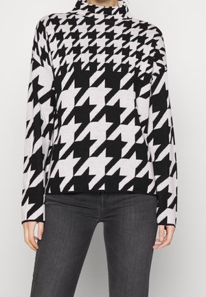 Haut à manches longues à motif pied-de-poule en noir et blanc avec un col montant, porté avec un pantalon gris foncé.