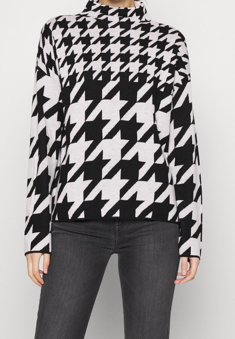 Haut à manches longues à motif pied-de-poule en noir et blanc avec un col montant, porté avec un pantalon gris foncé.