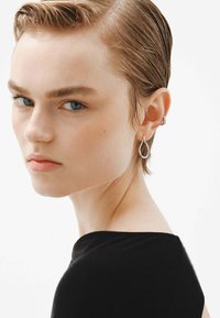 Pendientes plateados en forma de lágrima cuelgan de una oreja, complementando un retrato en primer plano de una persona con cabello corto y una prenda negra.
