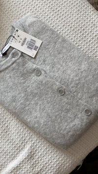 Grå stickad tröja liggande platt med fem knappar framtill. Materialet verkar mjukt och texturerat. Fastsydd etikett visar storlek M.