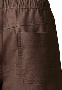 Gros plan sur un pantalon en tissu marron avec une ceinture élastique et une grande poche rectangulaire cousue sur le côté.