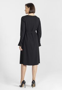 Czarna sukienka z długimi rękawami, wcięciem w talii i rozkloszowanym dołem. Gładki materiał, okrągły dekolt i zamek z tyłu. Noszona z czarnymi szpilkami.