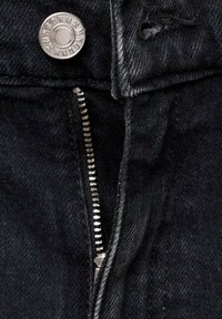 Detailní záběr na černou džínovinu s kovovým zipem a knoflíkem. Součástí designu je nenápadné otevírání zipu na jedné straně.