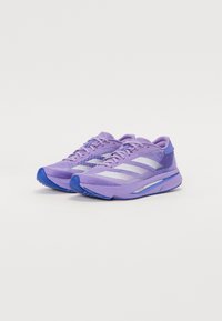 Zapatillas deportivas de color púrpura claro con una parte superior de malla texturizada, tres franjas blancas a los lados y acentos azules. Cuentan con una suela acolchada.