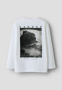 Witte lange mouw t-shirt met een zwart-wit afdruk van een kustlandschap, met tekst erboven en eronder, die thema's van keuze en emoties benadrukt.