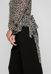 Blouse noire transparente avec imprimé léopard blanc, présentant des poignets volants et un détail de nouage sur le côté. Portée avec un pantalon noir.