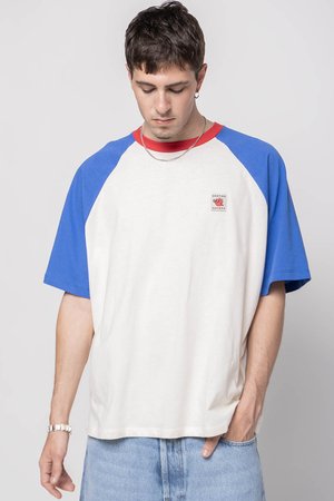 T-shirt à manches courtes avec un corps crème, des manches raglan bleues et un col rouge. Présente un petit patch logo sur la poitrine gauche. Matériau en coton.