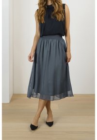 Femme portant un haut noir sans manches, une jupe grise mi-longue en tissu transparent, et des escarpins noirs à bout pointu, debout sur un sol en bois clair.