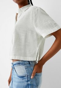 Biała lniana koszula z krótkim rękawem i kołnierzykiem, o luźnym krótkim kroju z bocznymi rozcięciami, noszona w połączeniu z jasnoniebieskimi dżinsami.