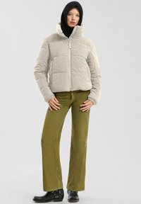 Helle graue gefütterte Jacke mit hohem Kragen, kombiniert mit olivgrünen Cordhosen und schwarzen Stiefeletten, die eine taillierte Silhouette aufweisen.