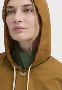 Veste à capuche marron en matériau lisse, avec une fermeture par boutons et un cordon de serrage crème. Superposée avec une chemise verte à col.