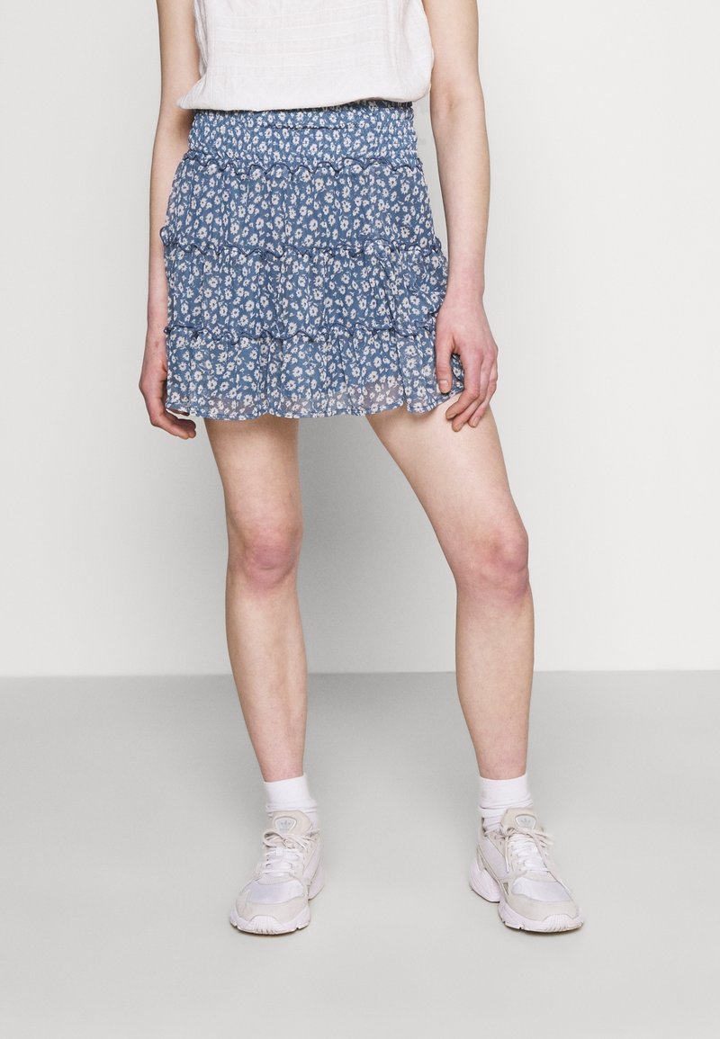 Personne portant une mini-jupe à volants bleue à motifs floraux, un haut blanc sans manches, des chaussettes blanches et des baskets blanches, debout devant un fond uni.