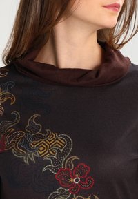T-shirt à manches longues avec un col roulé marron. Le tissu sombre présente des motifs floraux et géométriques colorés, avec une texture lisse et un léger éclat.