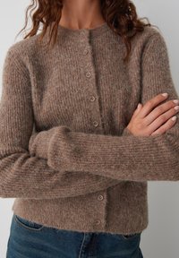 Bruine, geprofileerde cardigan gemaakt van een zachte stof met een ronde halslijn, vijf knopen aan de voorkant en aansluitende manchetten. Draagt over blauwe jeans.