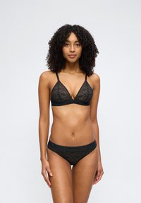 Γυναίκα με κατσαρά μαλλιά που φοράει μαύρο δαντελένιο μπραλέ και αντίστοιχα δαντελένια εσώρουχα, στέκεται μπροστά σε ένα απλό φωτεινό φόντο.