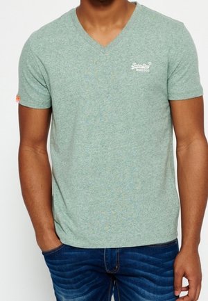Man draagt een lichtgroen T-shirt met V-hals en korte mouwen met een klein wit logo op de borst, gecombineerd met een blauwe spijkerbroek, handen ontspannen langs de zij.