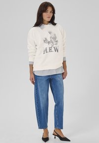 Felpa crema con grafica di un'aquila e testo "M.E.W.", sovrapposta a una camicia a righe. Jeans a vita alta blu e décolleté nere.