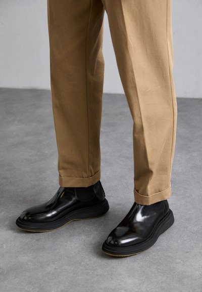 Botines negros brillantes de tobillo con paneles laterales elásticos, suela gruesa y costuras mínimas, combinados con pantalones beige con puños.