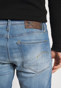 Siniset farkut kuluneella ilmeellä, joissa on ruskea nahkainen vyötärönauhan merkki, keltaiset ompeleet ja kaksi takataskua, joissa on kaarevat yksityiskohdat.