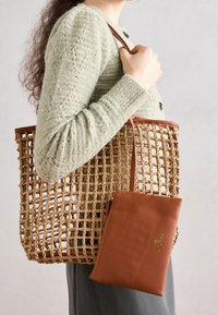 Bolsa de mano de paja trenzada con detalles en cuero marrón, que cuenta con un pouch interior desmontable de cuero marrón con un logo dorado.