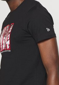 Černé tričko s krátkým rukávem má velký červený a bílý grafický potisk se nápisem "NBA" a malé bílé logo na levém rukávu.