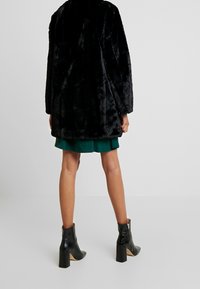 Svart fuskpälsjacka med lös passform och vida ärmar, i kombination med en grön kjol och svarta ankelboots med texturerade detaljer och staplade klackar.