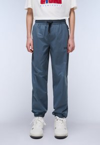 Pantalons de jogging légers et résistants à l'eau, de couleur bleue, avec des poignets élastiques, une taille à cordon de serrage et un logo sur le côté. Baskets blanches portées.