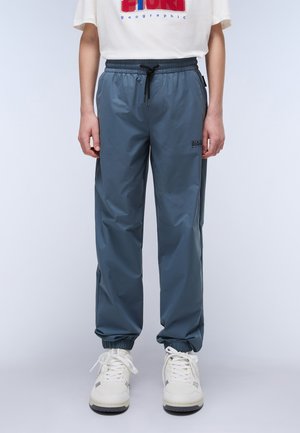 Pantalones de chándal ligeros y resistentes al agua, de color azul, con puños elásticos, cintura con cordón y detalle del logo en el lateral. Se llevan zapatillas blancas.