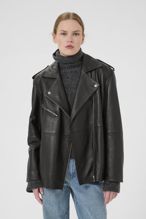 Veste de motard en cuir noir avec des revers surdimensionnés, des poches zippées angulaires et une silhouette carrée. Portée par-dessus un col roulé gris.