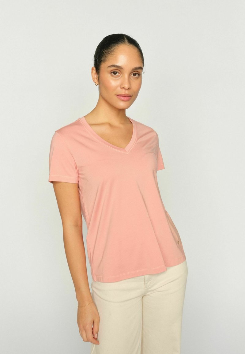 Haját hátrahúzott nő, világos rózsaszín V-nyakú pólót és törtfehér nadrágot visel, egyszerű világos háttér előtt áll.