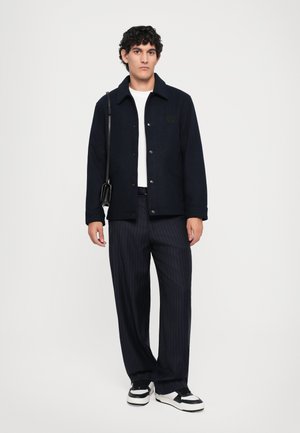 Navy wool blend jacket with a button closure and chest logo, paired with pinstriped trousers and black and white sneakers.