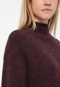Bordowy sweter z miękkiego, strukturalnego materiału dzianinowego, z wysokim kołnierzem i luźnym krojem, z prążkowanymi detalami i subtelnymi wariacjami kolorystycznymi.