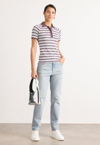 Gestreepte polo in bruin en blauw, gecombineerd met lichtblauwe jeans. Het model houdt een glanzende zilveren handtas vast. Witte sneakers maken de outfit compleet.