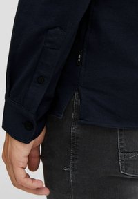 Primer plano de una mano de una persona a su lado, vistiendo una camiseta de manga larga azul marino con puños abotonados y jeans oscuros.