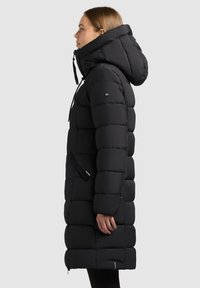 Schwarzer Steppmantel mit langem Schnitt, großer Kapuze und markanten gesteppte Partien. Verfügt über verstellbare Kordelzüge und seitliche Taschen.