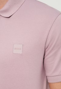 Ροζ ανοιχτό πουκάμισο πόλο σε υφασμάτινη υφή με γιακά· διαθέτει ένα μικρό, ανάγλυφο τετράγωνο λογότυπο «BOSS» στο στήθος.