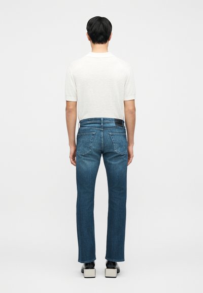 Τζιν παντελόνι σε στενή γραμμή με κλασική κοπή, που διαθέτει δύο πίσω τσέπες και μαύρο δερμάτινο ταμπελάκι στη μέση, σε συνδυασμό με ένα ανοιχτόχρωμο πουκάμισο.