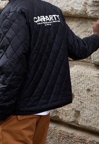 Černá prošívaná bunda s viditelným logem "CARHARTT" na zádech. Má volný střih a texturovanou tkaninu. Natažená přes hnědé kalhoty.